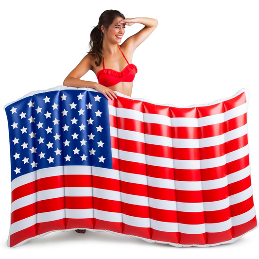 A smiling woman in a red bikini poses behind a large inflatable pool float designed to resemble the American flag, with red and white stripes and a blue field with white stars. She is looking into the distance with her hand above her eyes in a playful, patriotic pose. The background is white.