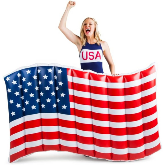 A woman in a USA swimsuit cheers with her fist raised behind a large inflatable pool float designed like the American flag.