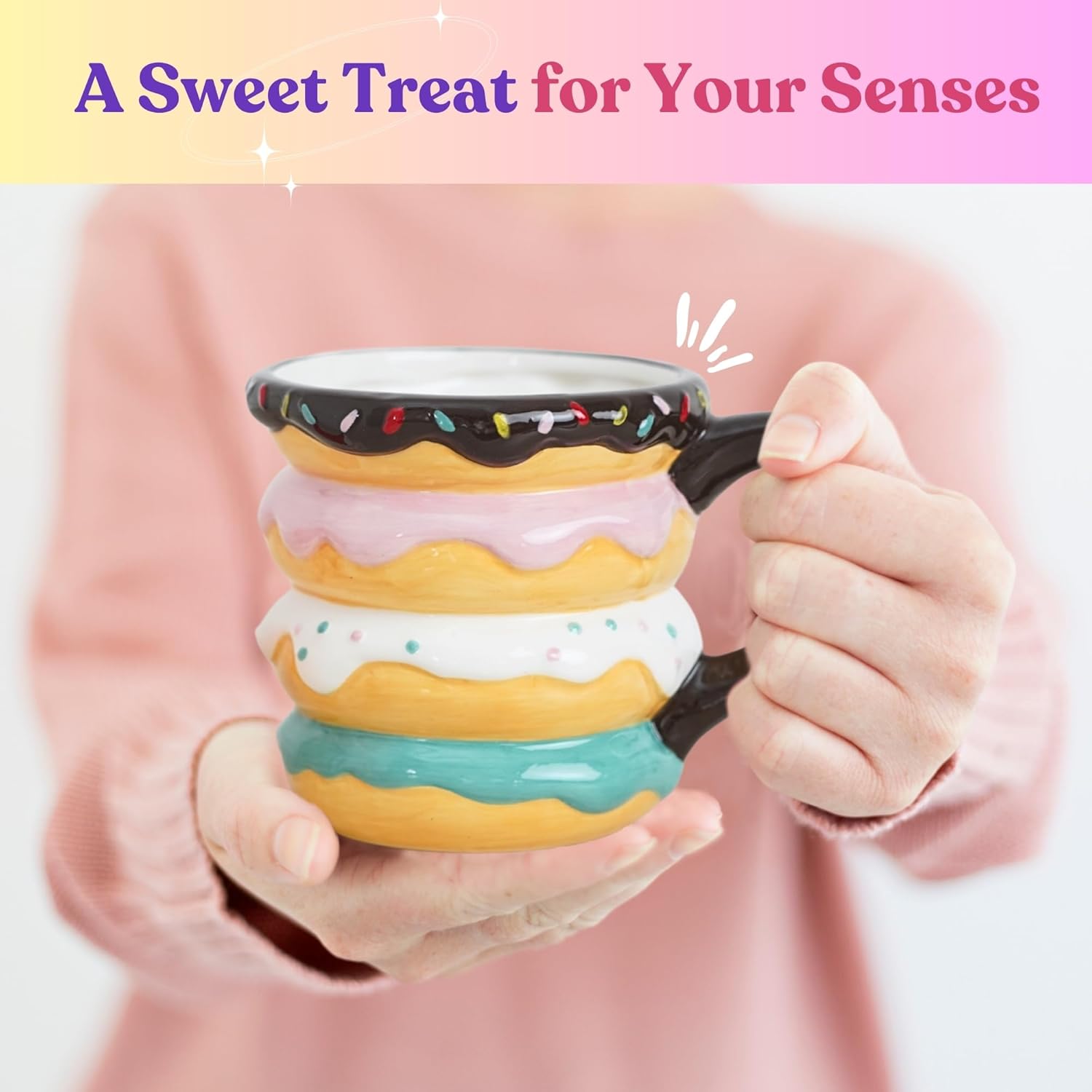 A person in a pink sweater holding a colorful ceramic donut-shaped mug with a sprinkle design, featuring layers of frosting in pastel colors.