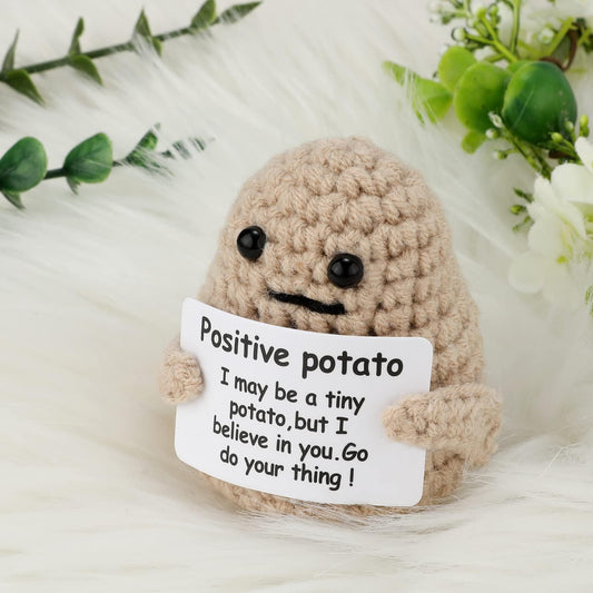 A small, crocheted plush toy shaped like a potato with black button eyes and a simple black stitched mouth. The plush toy is holding a white card with a motivational message that reads: "Positive potato. I may be a tiny potato, but I believe in you. Go do your thing!" The toy has short arms and a round, soft appearance, giving it a cute and encouraging vibe.